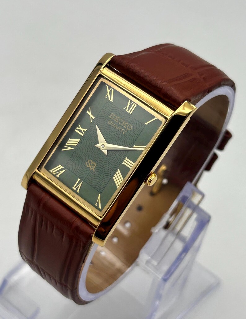 Puede incluir: Un reloj Seiko Quartz rectangular con caja dorada y esfera verde con n&uacute;meros romanos. El reloj tiene una correa de cuero marr&oacute;n. El reloj se muestra sobre un soporte transparente.