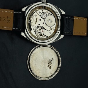 May include: A vintage Tudor watch with the back removed, showcasing the internal mechanisms. The watch features a silver case and a black leather strap. The watch face is absent, and the watch back is positioned beside the watch.