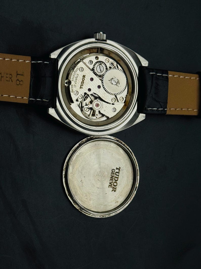 May include: A vintage Tudor watch with the back removed, showcasing the internal mechanisms. The watch features a silver case and a black leather strap. The watch face is absent, and the watch back is positioned beside the watch.