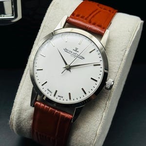 Puede incluir: Un reloj plateado con esfera blanca y agujas negras, con la inscripci&oacute;n "JAEGER-LECOULTRE CLUB" y "SWISS MADE". Tiene una correa de cuero marr&oacute;n.