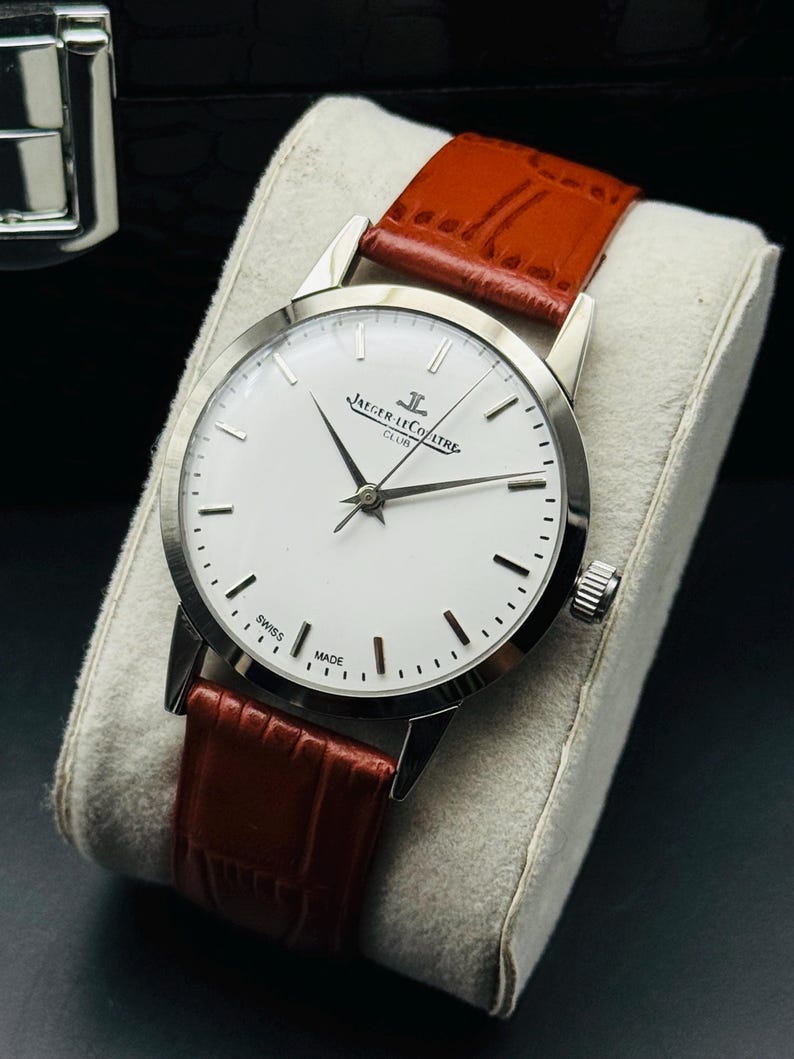 Puede incluir: Un reloj plateado con esfera blanca y agujas negras, con la inscripci&oacute;n "JAEGER-LECOULTRE CLUB" y "SWISS MADE". Tiene una correa de cuero marr&oacute;n.