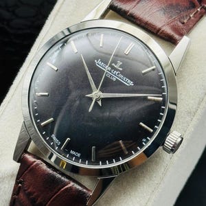 May include: A close-up of a Jaeger-LeCoultre Club watch with a dark face and silver hands. The watch has a brown leather band. The words "SWISS MADE" are visible on the watch face.
