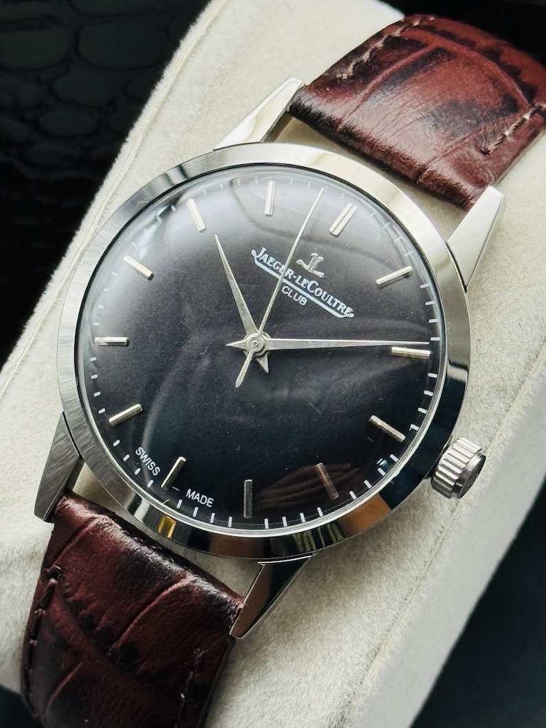 May include: A close-up of a Jaeger-LeCoultre Club watch with a dark face and silver hands. The watch has a brown leather band. The words "SWISS MADE" are visible on the watch face.
