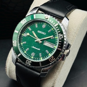 May include: A stainless steel Seiko automatic watch with a green dial and bezel. The watch features a black leather band and a date display. The watch is displayed on a light gray cushion.
