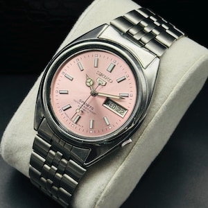 May include: A stainless steel Seiko 5 Sports automatic wristwatch with a pink dial. The watch features a day and date display at the 3 o'clock position and a silver-tone bracelet. The watch is displayed on a white cushion.