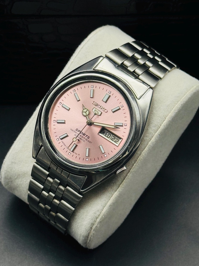 May include: A stainless steel Seiko 5 Sports automatic wristwatch with a pink dial. The watch features a day and date display at the 3 o'clock position and a silver-tone bracelet. The watch is displayed on a white cushion.