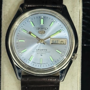 May include: A silver-toned Seiko 5 Sports automatic watch with a light blue dial and a brown leather strap. The watch face displays the day and date, with luminous hands and markers. The watch is marked "Japan 7009 676L R".