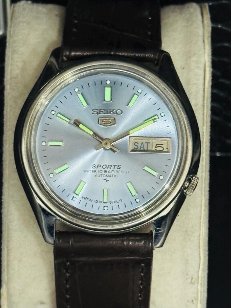 May include: A silver-toned Seiko 5 Sports automatic watch with a light blue dial and a brown leather strap. The watch face displays the day and date, with luminous hands and markers. The watch is marked "Japan 7009 676L R".