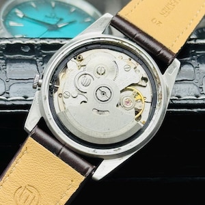 May include: Close-up of a silver wristwatch with a brown leather strap. The watch's back is open, revealing the intricate internal mechanisms, including gears and a balance wheel. The watch has a light-colored leather strap.