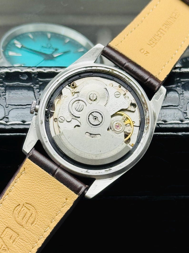 May include: Close-up of a silver wristwatch with a brown leather strap. The watch's back is open, revealing the intricate internal mechanisms, including gears and a balance wheel. The watch has a light-colored leather strap.