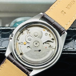 May include: Close-up of a silver-toned wristwatch with a brown leather strap. The watch's intricate internal mechanism is visible, featuring gears, jewels, and the text "SEIKO TWENTY-ONE JEWELS". The strap is marked "LEATHER".
