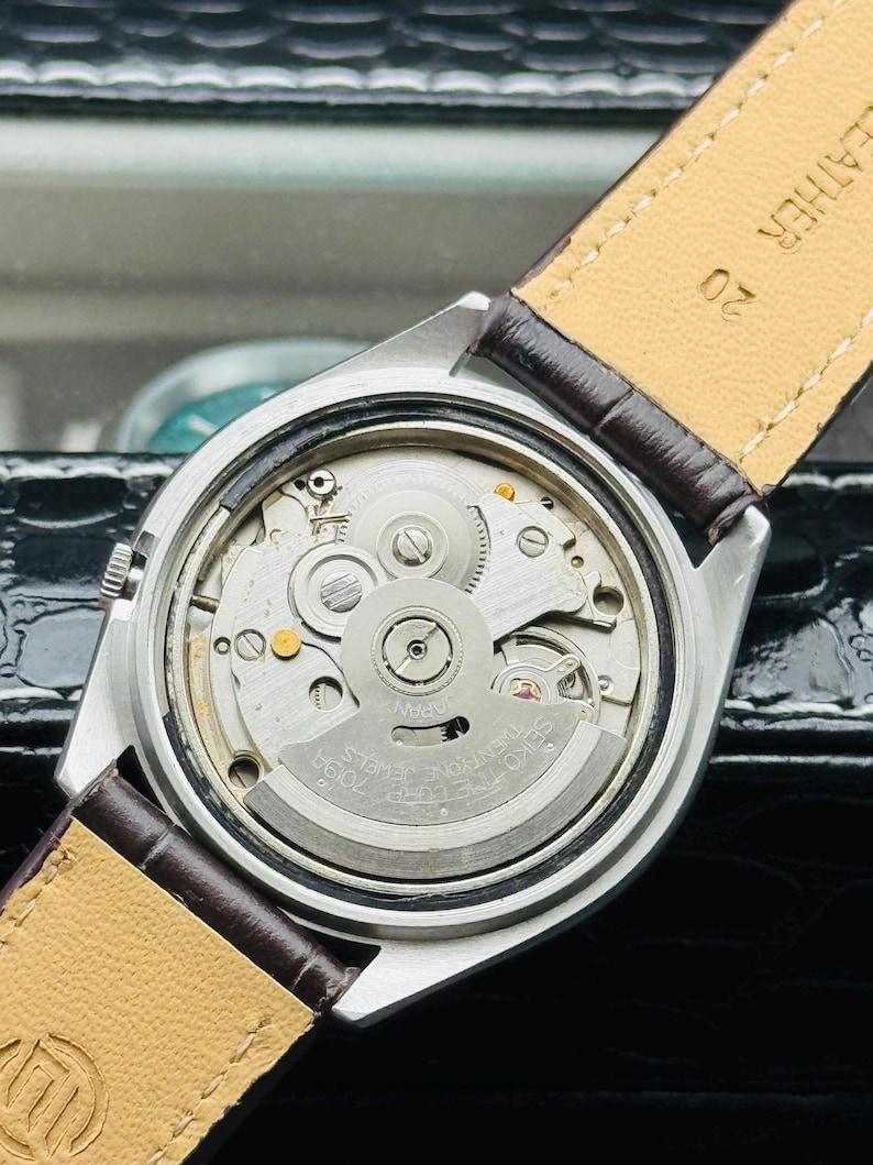 May include: Close-up of a silver-toned wristwatch with a brown leather strap. The watch's intricate internal mechanism is visible, featuring gears, jewels, and the text "SEIKO TWENTY-ONE JEWELS". The strap is marked "LEATHER".