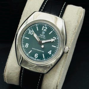 May include: A vintage Tudor Oyster watch with a green face and silver casing. The watch has a black leather strap with white stitching. The dial shows the Tudor logo, hour markers, and the words "SHOCK RESISTING".