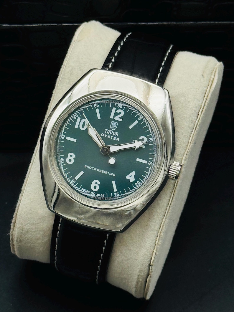 May include: A vintage Tudor Oyster watch with a green face and silver casing. The watch has a black leather strap with white stitching. The dial shows the Tudor logo, hour markers, and the words "SHOCK RESISTING".