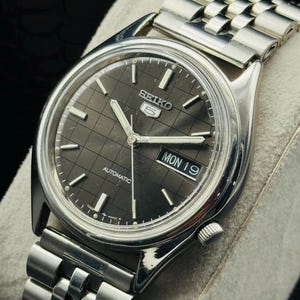 Op de afbeelding: Een zilverkleurig Seiko 5 automatisch horloge met een donkergrijze geruite wijzerplaat. Het horloge heeft een dag- en datumweergave, zilveren wijzers en een roestvrijstalen band. Het horloge wordt gepresenteerd op een lichtgrijze ondergrond.