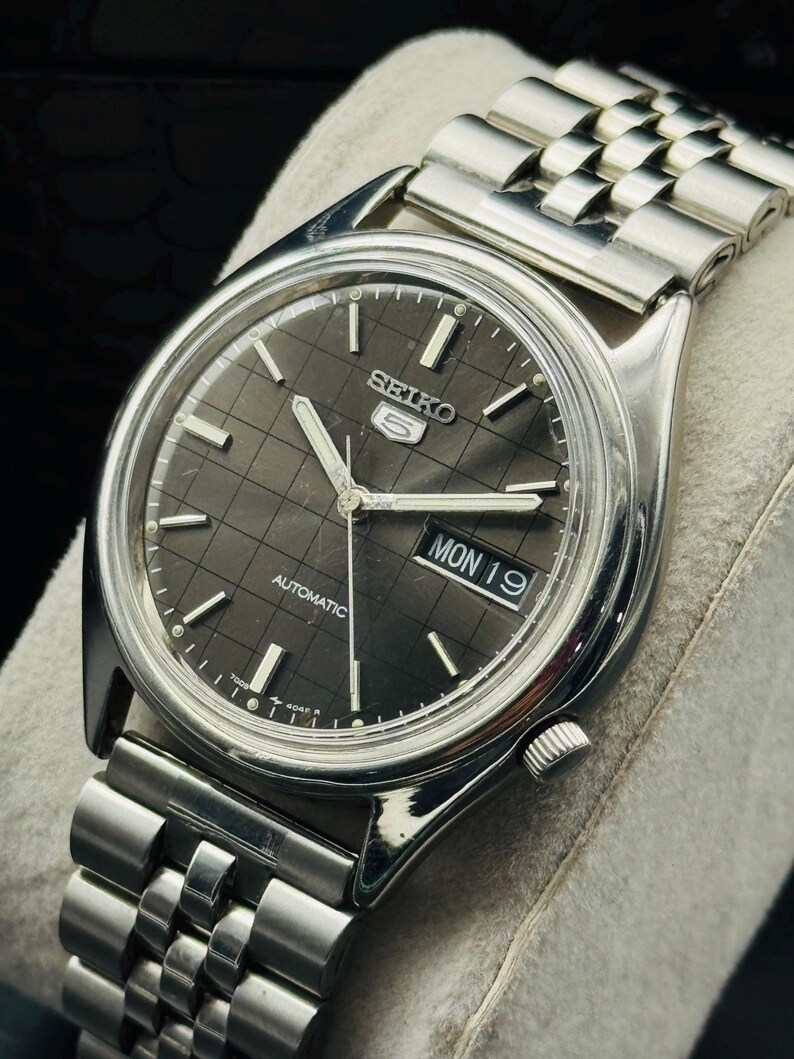 Op de afbeelding: Een zilverkleurig Seiko 5 automatisch horloge met een donkergrijze geruite wijzerplaat. Het horloge heeft een dag- en datumweergave, zilveren wijzers en een roestvrijstalen band. Het horloge wordt gepresenteerd op een lichtgrijze ondergrond.