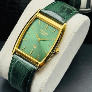 Könnte beinhalten: Eine goldfarbene Seiko Quartz-Uhr mit einem rechteckigen grünen Zifferblatt und goldenen Zeigern. Die Uhr hat ein grünes Lederarmband. Die Uhr wird auf einem weißen Kissen präsentiert.