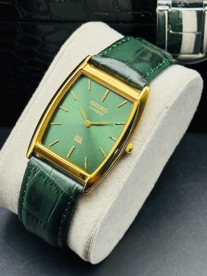 May include: A gold-toned Seiko Quartz watch with a rectangular green face and gold hands. The watch has a green leather band. The watch is displayed on a white cushion.