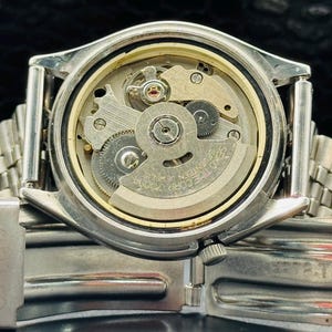 Op de afbeelding: Close-up van een zilverkleurig polshorloge met de achterkant verwijderd, waardoor het ingewikkelde mechanische uurwerk zichtbaar wordt. Het horloge heeft een zilveren lunette en een zilveren metalen band. De interne tandwielen en componenten zijn zichtbaar.