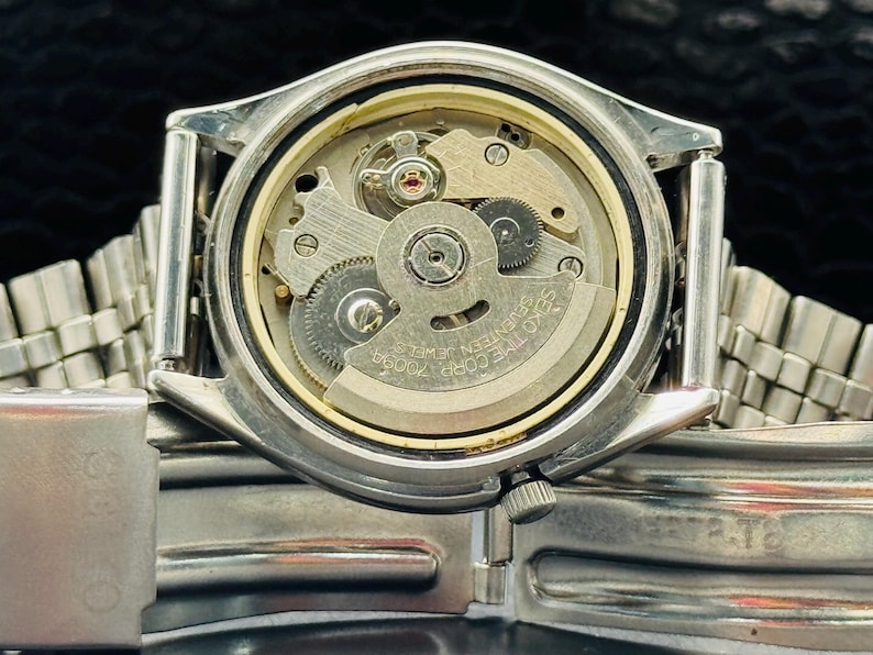 Op de afbeelding: Close-up van een zilverkleurig polshorloge met de achterkant verwijderd, waardoor het ingewikkelde mechanische uurwerk zichtbaar wordt. Het horloge heeft een zilveren lunette en een zilveren metalen band. De interne tandwielen en componenten zijn zichtbaar.