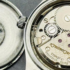 May include: Open view of a vintage wristwatch with the back cover detached. The watch mechanism is visible, showcasing intricate gears, jewels, and the "LeCoultre Co" inscription. The back cover displays "ACER INOXIDABLE" and "LeCoultre Co" text.