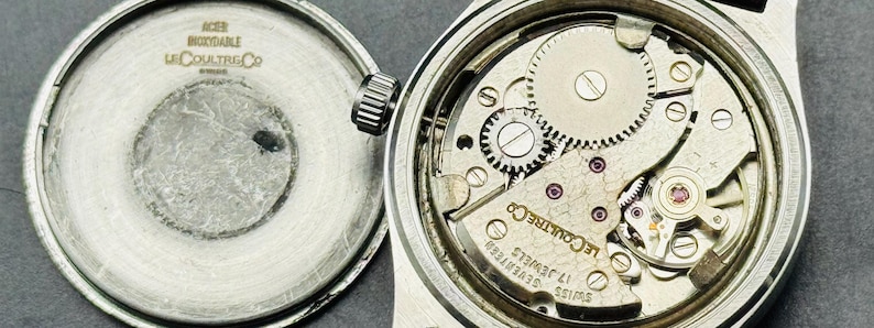 May include: Open view of a vintage wristwatch with the back cover detached. The watch mechanism is visible, showcasing intricate gears, jewels, and the "LeCoultre Co" inscription. The back cover displays "ACER INOXIDABLE" and "LeCoultre Co" text.