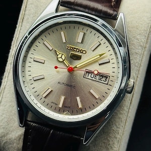 May include: A silver Seiko automatic wristwatch with a light gray face, gold hands and markers, and a brown leather strap. The watch face displays the day and date, with the text "AUTOMATIC" and "JAPAN MADE".