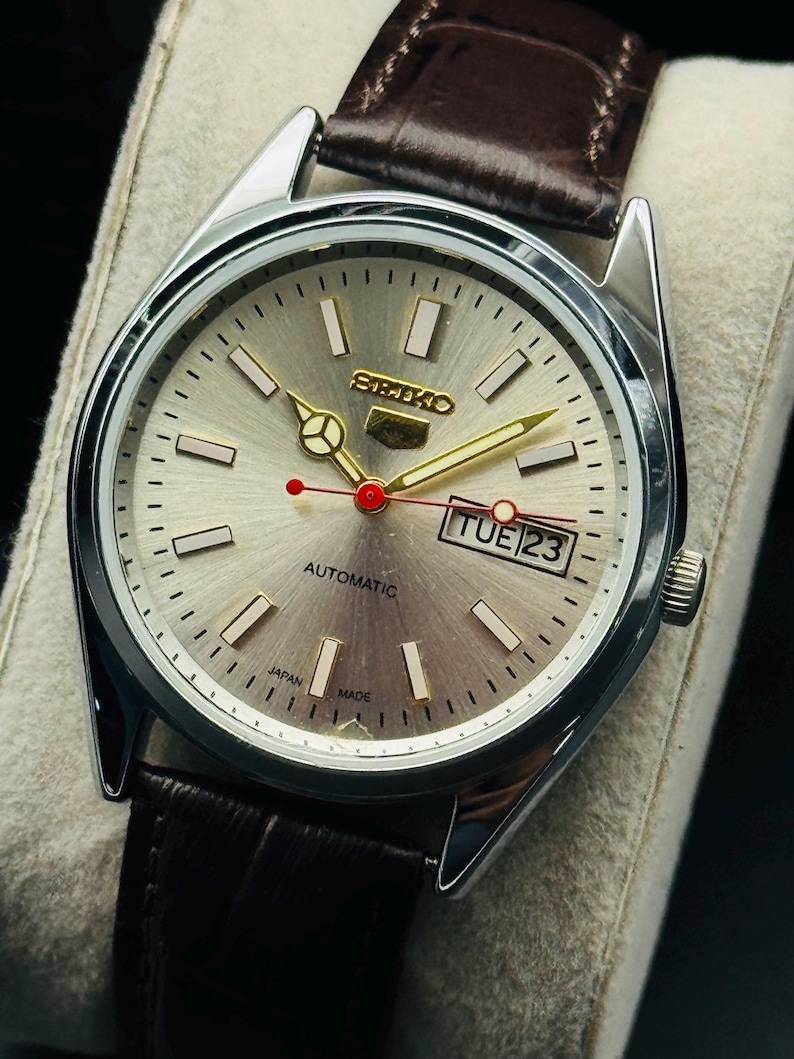 May include: A silver Seiko automatic wristwatch with a light gray face, gold hands and markers, and a brown leather strap. The watch face displays the day and date, with the text "AUTOMATIC" and "JAPAN MADE".