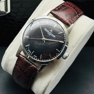 May include: A silver-toned wristwatch with a dark face and brown leather band. The watch face has silver hour markers and hands. The band is a rich, dark brown. The watch is displayed on a white cushion.