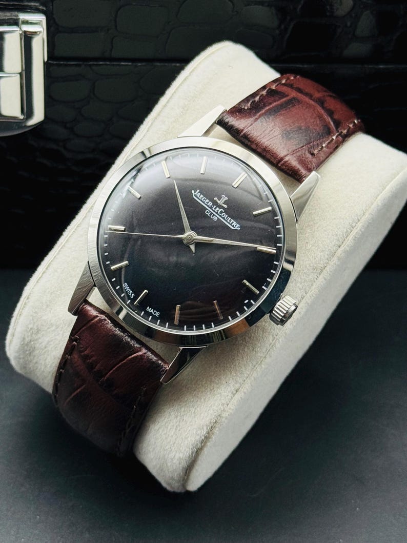 May include: A silver-toned wristwatch with a dark face and brown leather band. The watch face has silver hour markers and hands. The band is a rich, dark brown. The watch is displayed on a white cushion.