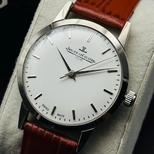 Puede incluir: Un reloj Jaeger-LeCoultre Club plateado con esfera blanca y agujas negras. Tiene una correa de cuero roja. La esfera del reloj muestra el texto "JAEGER-LECOULTRE CLUB" y "SWISS MADE".