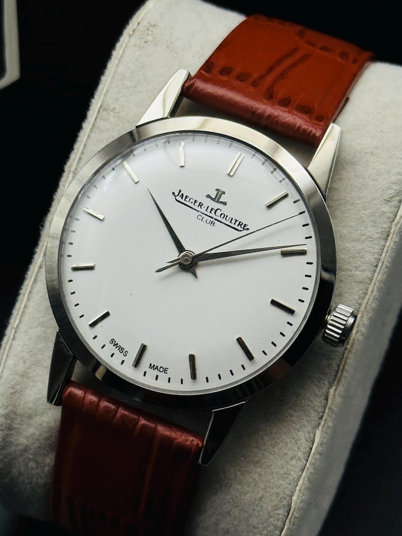 Puede incluir: Un reloj Jaeger-LeCoultre Club plateado con esfera blanca y agujas negras. Tiene una correa de cuero roja. La esfera del reloj muestra el texto "JAEGER-LECOULTRE CLUB" y "SWISS MADE".