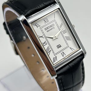 Puede incluir: Un reloj Seiko Quartz rectangular con caja plateada y correa de cuero negro. La esfera es blanca con n&uacute;meros romanos y agujas negras. La correa es de cuero negro con un patr&oacute;n de cocodrilo.