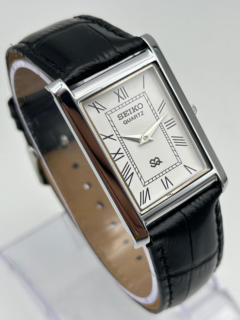 Puede incluir: Un reloj Seiko Quartz rectangular con caja plateada y correa de cuero negro. La esfera es blanca con n&uacute;meros romanos y agujas negras. La correa es de cuero negro con un patr&oacute;n de cocodrilo.