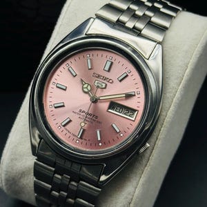May include: A stainless steel Seiko 5 Sports automatic watch with a pink dial. The watch features a day and date display, luminous hands and markers, and a stainless steel bracelet. The text on the dial reads "SPORTS WATER 10 BAR RESIST AUTOMATIC".