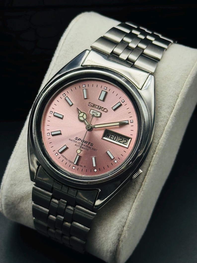 May include: A stainless steel Seiko 5 Sports automatic watch with a pink dial. The watch features a day and date display, luminous hands and markers, and a stainless steel bracelet. The text on the dial reads "SPORTS WATER 10 BAR RESIST AUTOMATIC".
