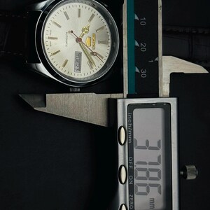 May include: Silver-toned Seiko automatic watch with a light-colored face, gold-colored hands, and a black leather strap. The watch is being measured with a digital caliper, displaying a measurement of 3.786 mm.