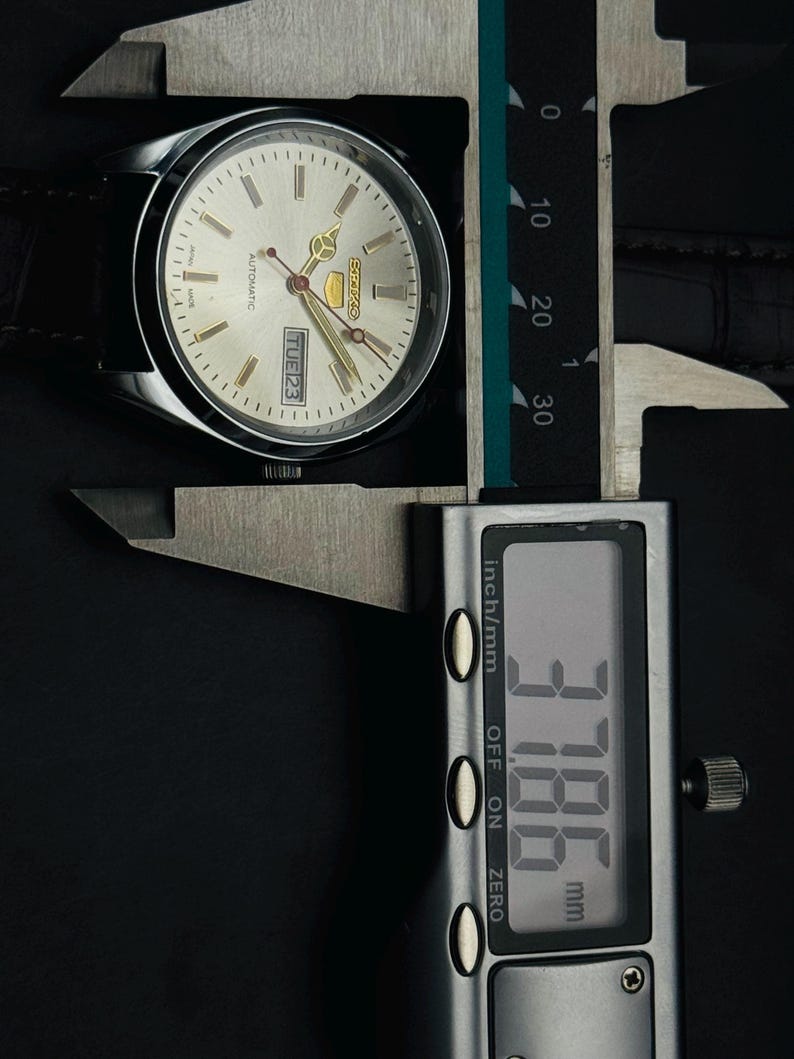 May include: Silver-toned Seiko automatic watch with a light-colored face, gold-colored hands, and a black leather strap. The watch is being measured with a digital caliper, displaying a measurement of 3.786 mm.