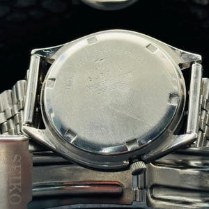 Op de afbeelding: Close-up van een zilverkleurig Seiko horloge. De achterkant van de horlogekast is zichtbaar, met gegraveerde tekst waaronder "Roestvrij staal" en "Waterbestendig". De horlogeband is een mesh-stijl en het Seiko-logo is zichtbaar aan de zijkant.