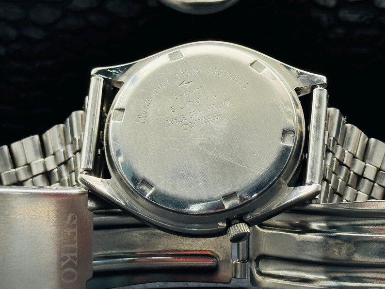 Op de afbeelding: Close-up van een zilverkleurig Seiko horloge. De achterkant van de horlogekast is zichtbaar, met gegraveerde tekst waaronder "Roestvrij staal" en "Waterbestendig". De horlogeband is een mesh-stijl en het Seiko-logo is zichtbaar aan de zijkant.