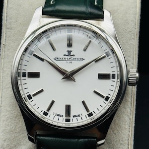 May include: A silver-toned Jaeger-LeCoultre Club watch with a white face and black markers. The watch has a green leather band. The watch face displays the Jaeger-LeCoultre logo and the words "CLUB", "T SWISS MADE T".
