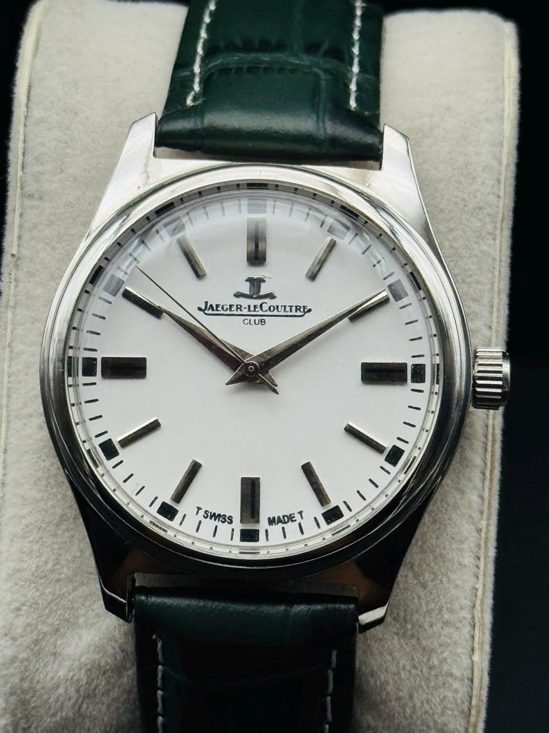 May include: A silver-toned Jaeger-LeCoultre Club watch with a white face and black markers. The watch has a green leather band. The watch face displays the Jaeger-LeCoultre logo and the words "CLUB", "T SWISS MADE T".