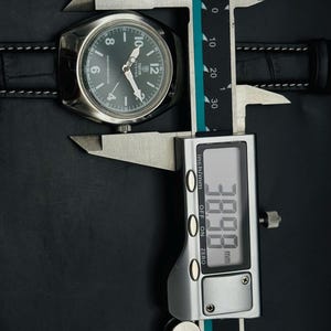 May include: A silver wristwatch with a dark green face and black leather strap is measured with a digital calliper. The calliper displays a measurement of 38.98 mm. The watch face has white hands and numerals.