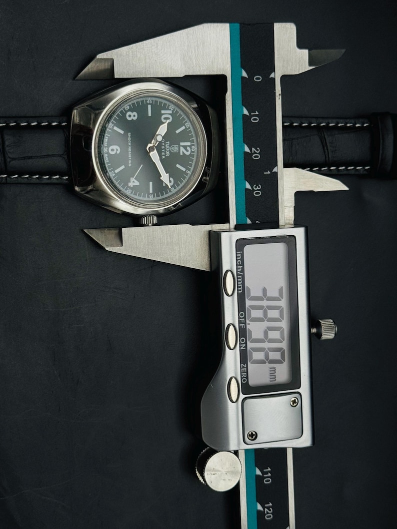 May include: A silver wristwatch with a dark green face and black leather strap is measured with a digital calliper. The calliper displays a measurement of 38.98 mm. The watch face has white hands and numerals.