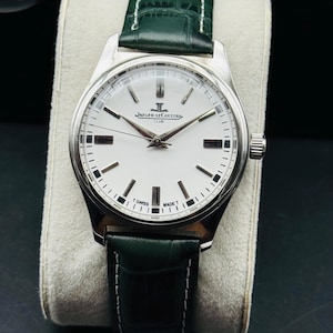 May include: A silver-toned Jaeger-LeCoultre Club watch with a white face, black markers, and a green leather strap. The watch face displays the Jaeger-LeCoultre logo and the words "SWISS MADE". The watch is displayed on a light-colored cushion.