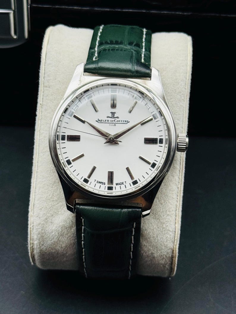 May include: A silver-toned Jaeger-LeCoultre Club watch with a white face, black markers, and a green leather strap. The watch face displays the Jaeger-LeCoultre logo and the words "SWISS MADE". The watch is displayed on a light-colored cushion.