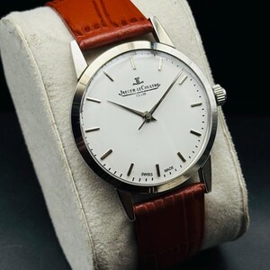 Puede incluir: Un reloj plateado con esfera blanca y correa de cuero marr&oacute;n. El reloj presenta el texto "JAEGER-LECOULTRE CLUB" y "SWISS MADE". El reloj se exhibe sobre un coj&iacute;n blanco.
