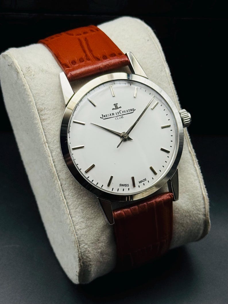 Puede incluir: Un reloj plateado con esfera blanca y correa de cuero marr&oacute;n. El reloj presenta el texto "JAEGER-LECOULTRE CLUB" y "SWISS MADE". El reloj se exhibe sobre un coj&iacute;n blanco.