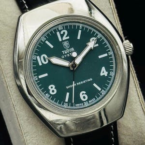May include: A vintage Tudor Oyster watch with a dark green face, silver case, and black leather strap. The watch face features white numerals, hands, and the text "SHOCK RESISTING". The watch is displayed on a white surface.