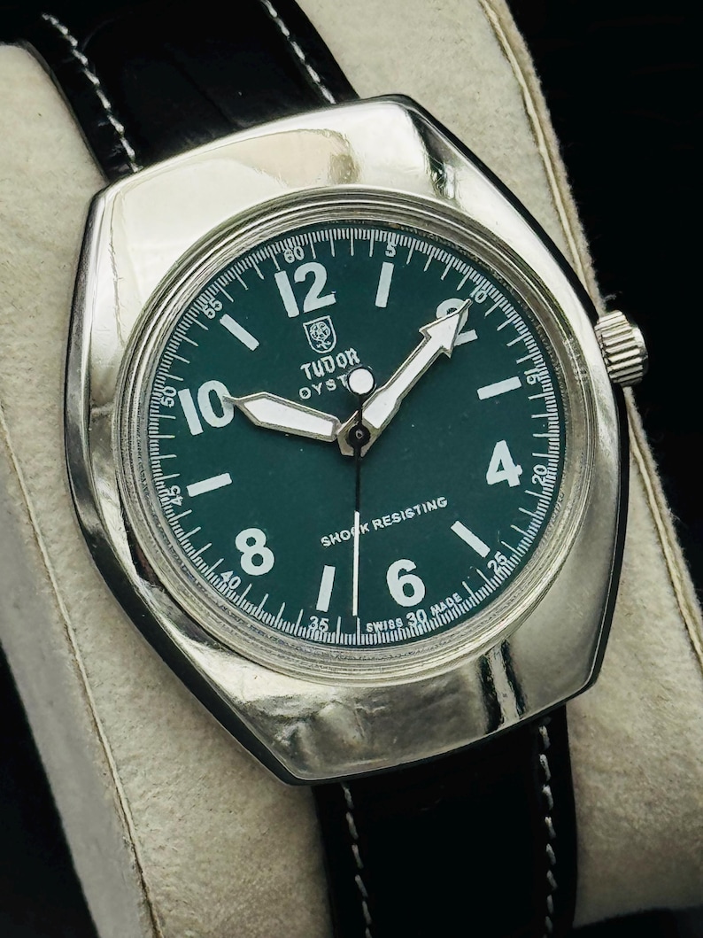 May include: A vintage Tudor Oyster watch with a dark green face, silver case, and black leather strap. The watch face features white numerals, hands, and the text "SHOCK RESISTING". The watch is displayed on a white surface.